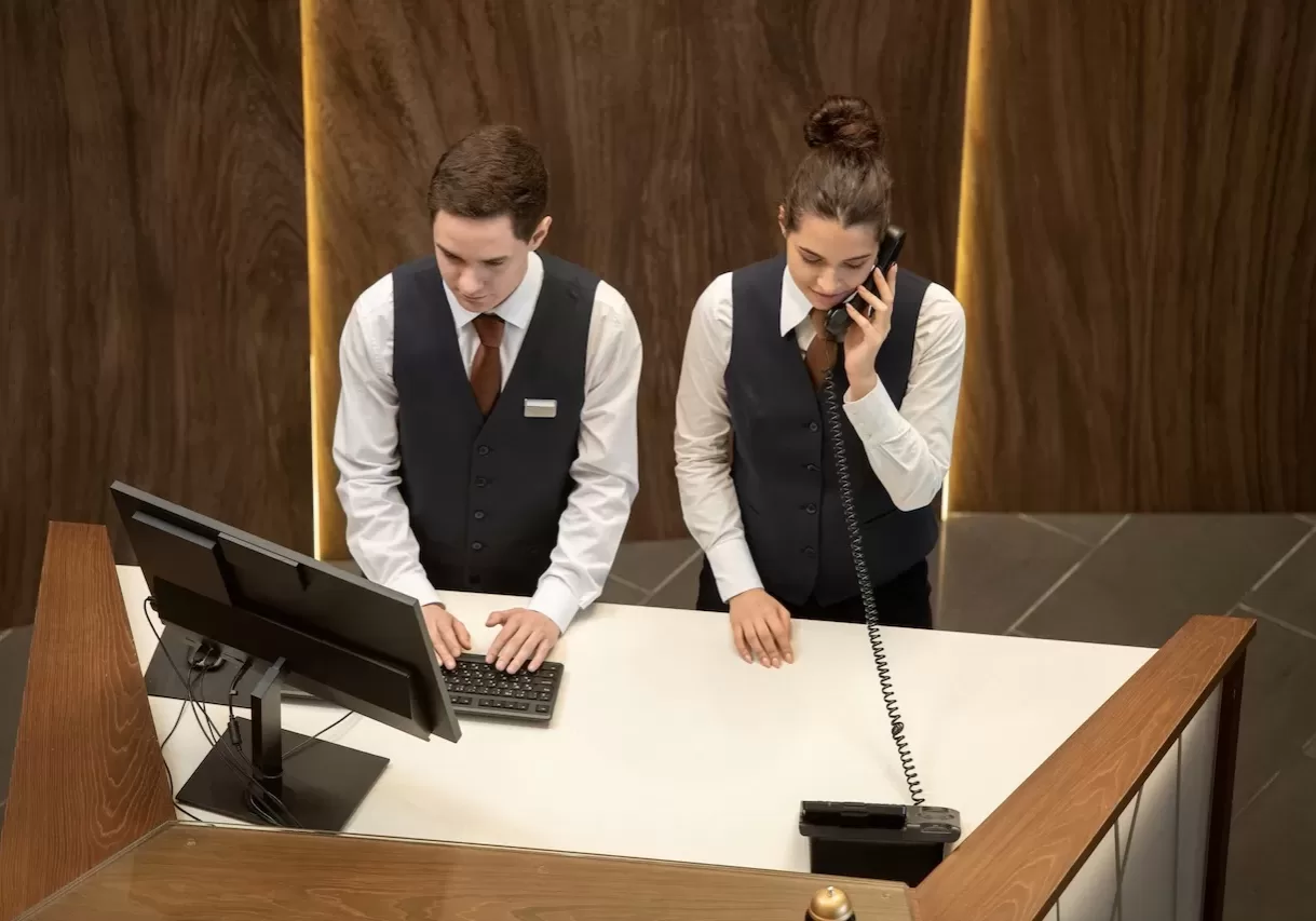 One of two young hotel receptionists standing by counter and looking at computer screen while his colleague answering phone near by